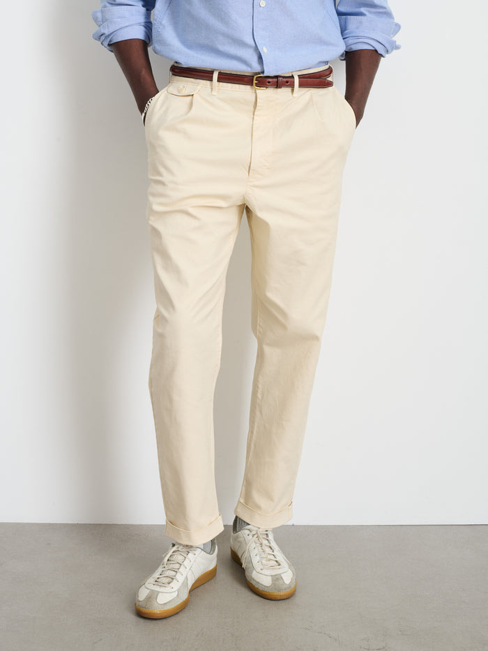 Wearing a light blue shirt, Standard Pleated Pant in Chino, brown belt, and white sneakers, a person stands against a white wall with hands in pockets. Their face is not visible.