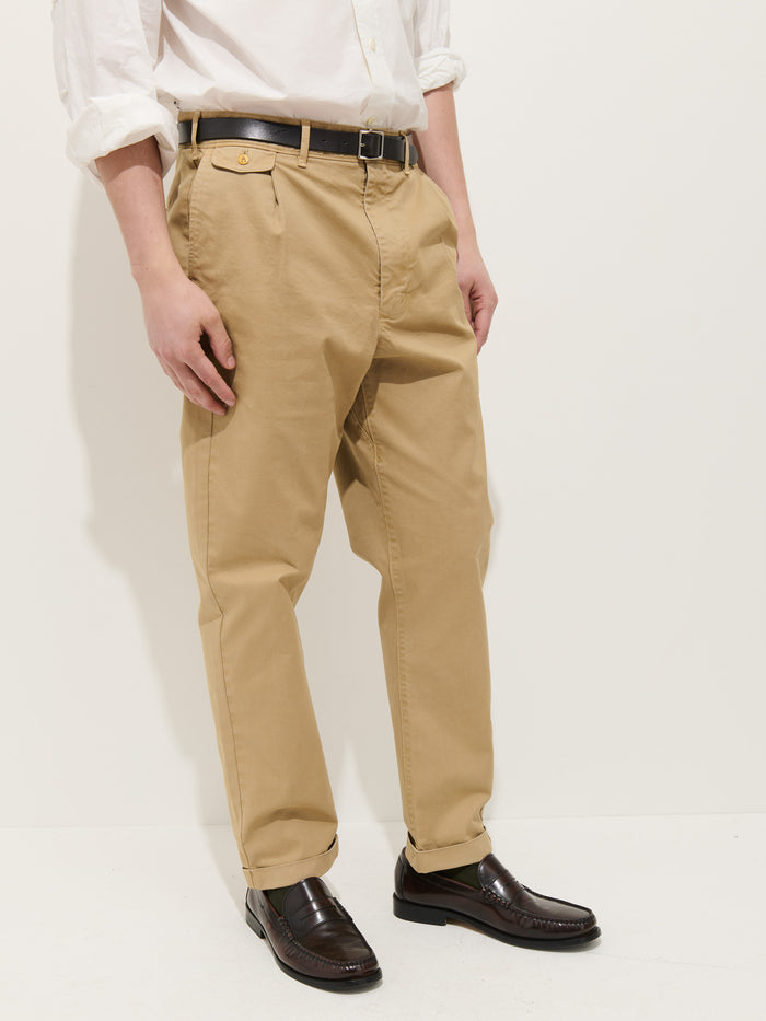 A model stands against a plain white background wearing the Standard Pleated Pant in Chino—beige cotton twill, relaxed fit—with a white long-sleeve shirt, black belt, and brown loafers. Their hands are at their sides and their face is not visible.
