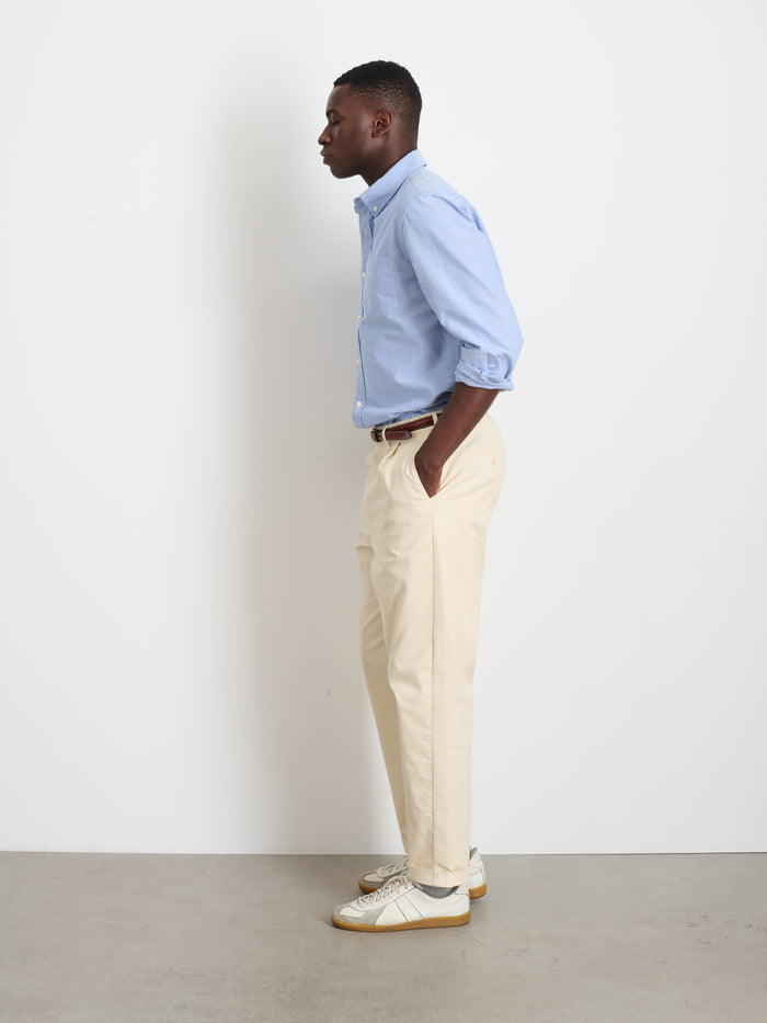 A man in profile stands against a white wall, wearing a light blue button-down shirt, the Standard Pleated Pant in Chino, a brown belt, and white sneakers. His hands are in his pockets as he looks straight ahead.