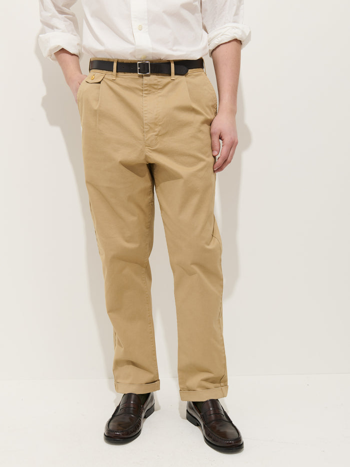 A person wearing the Standard Pleated Pant in Chino, a white shirt, black belt, and black loafers stands against a plain white background with hands in their pockets. Their face is not shown.