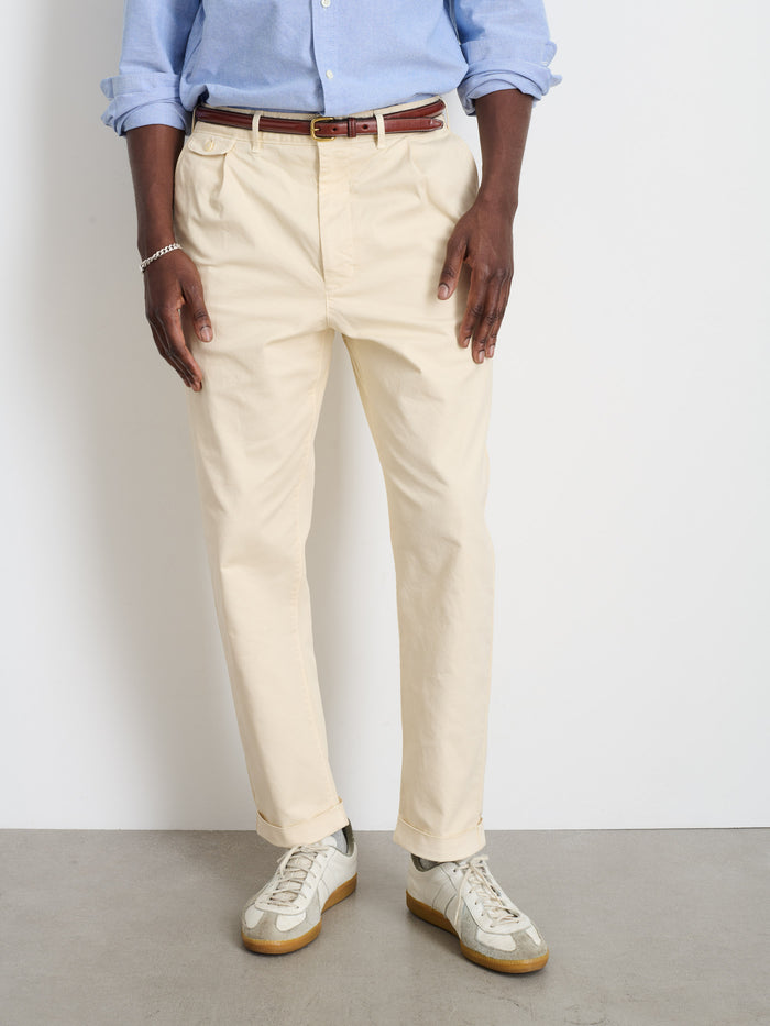 Only the lower torso and legs are visible, wearing Standard Pleated Pant in Chino (beige cotton twill), paired with a relaxed fit light blue shirt, brown belt, and white sneakers with gum soles against a white wall.
