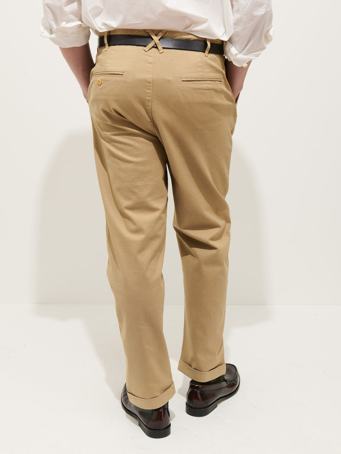 A person wearing the Standard Pleated Pant in Chino, a white long-sleeve shirt with rolled-up sleeves, a black belt, and dark brown dress shoes stands with their back to the camera against a plain white background.