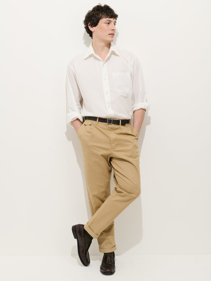 A person wearing a white long-sleeve button-up shirt, Standard Pleated Pant in Chino, and dark shoes stands against a plain white background with hands in pockets and one leg crossed over the other.