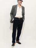 A man wearing sunglasses, a grey blazer, a cream 100% cotton button-up shirt, the Standard Pleated Pant In Cord in dark navy, black loafers, and orange socks stands against a plain white background with his hands in his pockets.