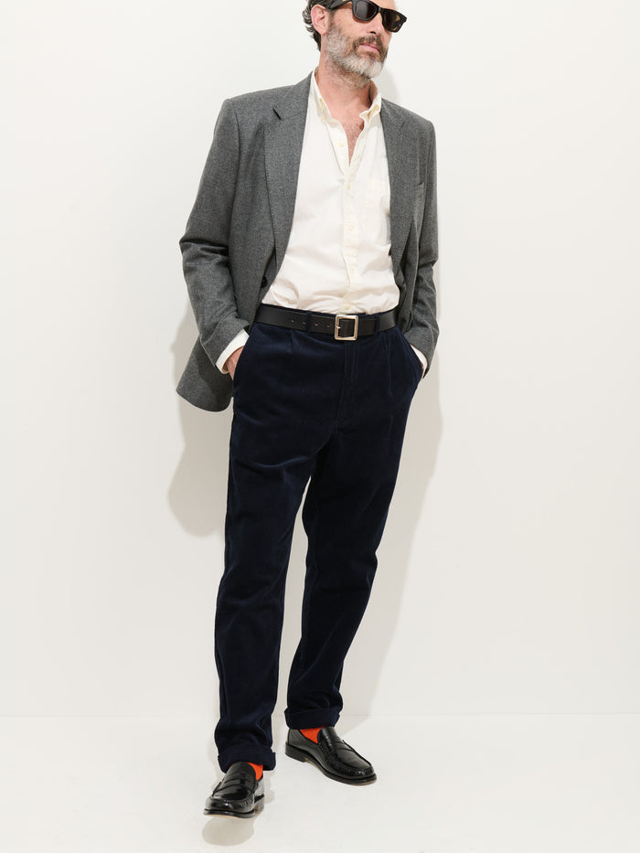 A man wearing sunglasses, a grey blazer, a cream 100% cotton button-up shirt, the Standard Pleated Pant In Cord in dark navy, black loafers, and orange socks stands against a plain white background with his hands in his pockets.