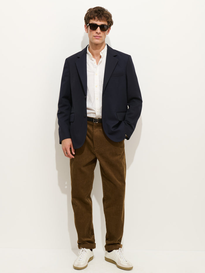 A man stands against a white background wearing sunglasses, a navy blazer with corozo buttons, a white button-up shirt, the Standard Pleated Pant In Cord in brown, and white sneakers.