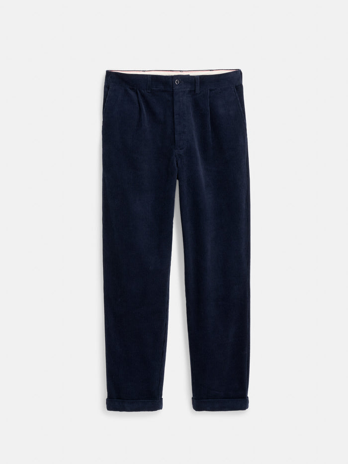 The Standard Pleated Pant In Cord, crafted in NYC from 100% navy blue cotton corduroy, features front pleats, a button and zip closure, and rolled-up cuffs. Shown laid flat on a white background.