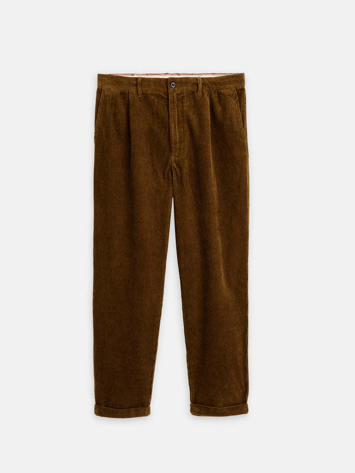 The Standard Pleated Pant In Cord: brown 100% cotton corduroy pants with corozo buttons, front pleats, and rolled-up cuffs, shown on a white background.