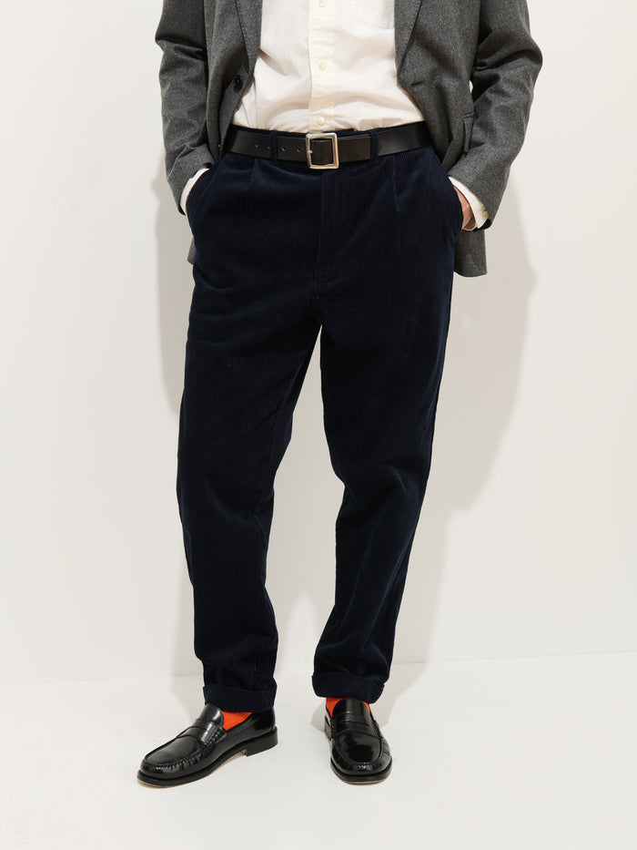 A person wearing a grey blazer, white shirt, black belt, orange socks, black loafers, and the Standard Pleated Pant In Cord (navy 100% cotton corduroy) stands with hands in pockets against a plain white background.