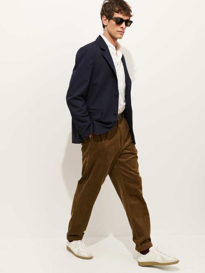 A person wearing sunglasses, a navy blazer with corozo buttons, a white shirt, Standard Pleated Pant In Cord in brown, and white sneakers walks against a plain white background.