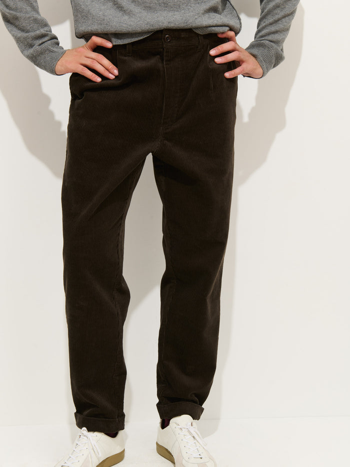 Wearing Standard Pleated Pant In Cord (dark brown, 100% cotton), a light gray long-sleeve top, and white sneakers, a person stands hands on hips against a white background. Only the lower body is visible; upper body and head are cropped out.