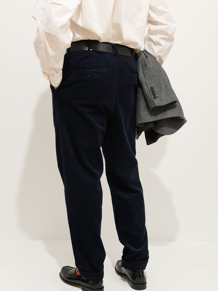 A person faces away, wearing the Standard Pleated Pant In Cord in dark blue, paired with a white shirt and black shoes. A gray blazer is draped over their left arm. The plain, light background highlights the NYC-made pants.