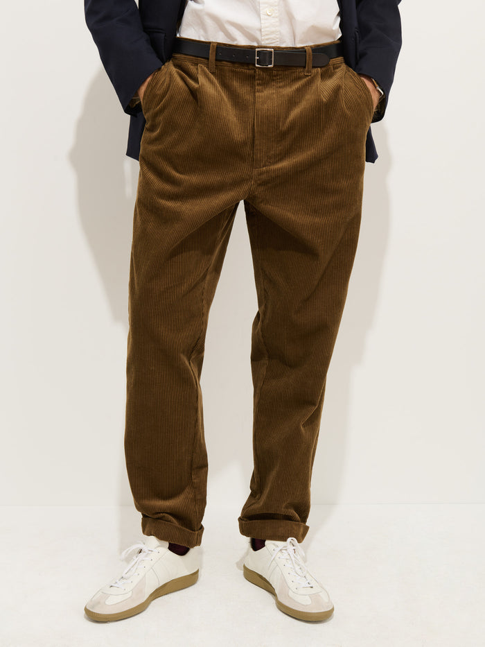 A person stands against a plain background wearing the Standard Pleated Pant In Cord (100% cotton, brown), a white shirt, black belt, dark blazer with corozo buttons, and white sneakers. Their hands are in their pockets and face is not visible.