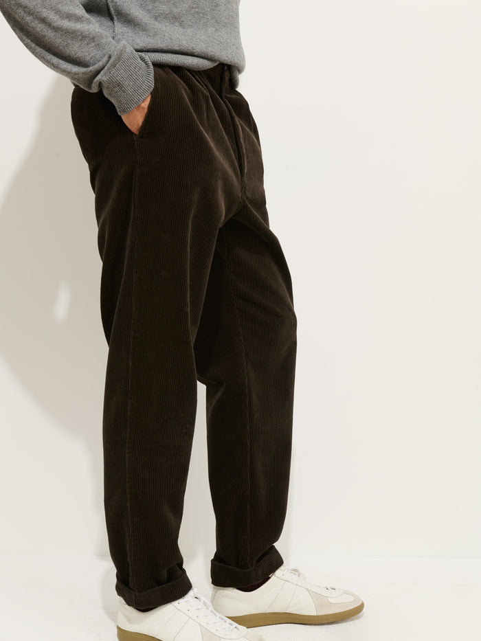 Wearing the Standard Pleated Pant In Cord (dark brown, 100% cotton), a gray sweater, and white sneakers, a person stands with one hand in their pocket against a plain white background.