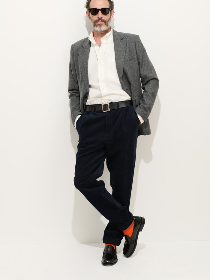 A man in a gray blazer, white shirt, and Standard Pleated Pant In Cord stands against a white background. He wears sunglasses, orange socks, black shoes, has hands in his pockets, and crosses one leg over the other.