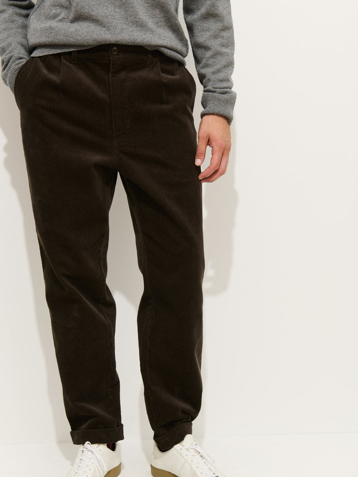 A person wearing a grey sweater, the Standard Pleated Pant In Cord in dark brown 100% cotton corduroy, and white sneakers stands against a plain white background with hands in pockets.