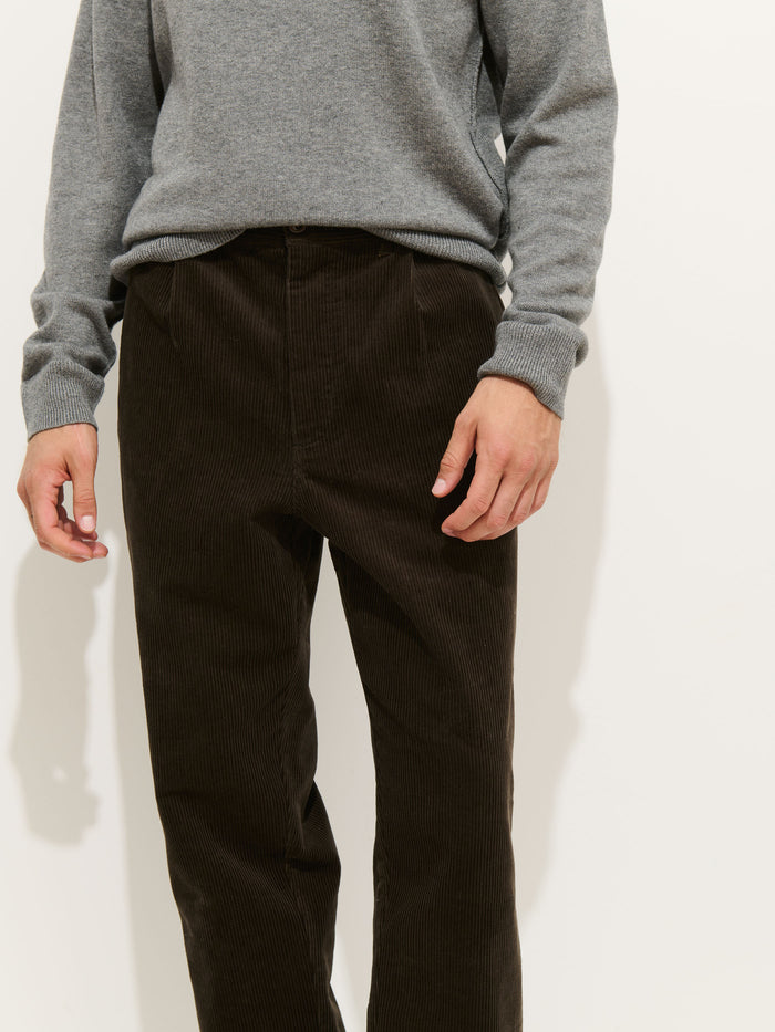 A person wears a light gray long-sleeve sweater tucked into high-waisted, dark brown Standard Pleated Pant in Cord made in NYC, standing against a plain white background. The individual's head is not visible in the image.