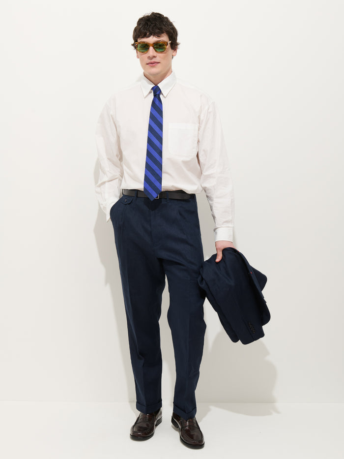 A man wearing sunglasses, a white dress shirt, a blue striped tie, brown shoes, and the Standard Pleated Pant In Linen stands holding a matching navy jacket against a plain white background.