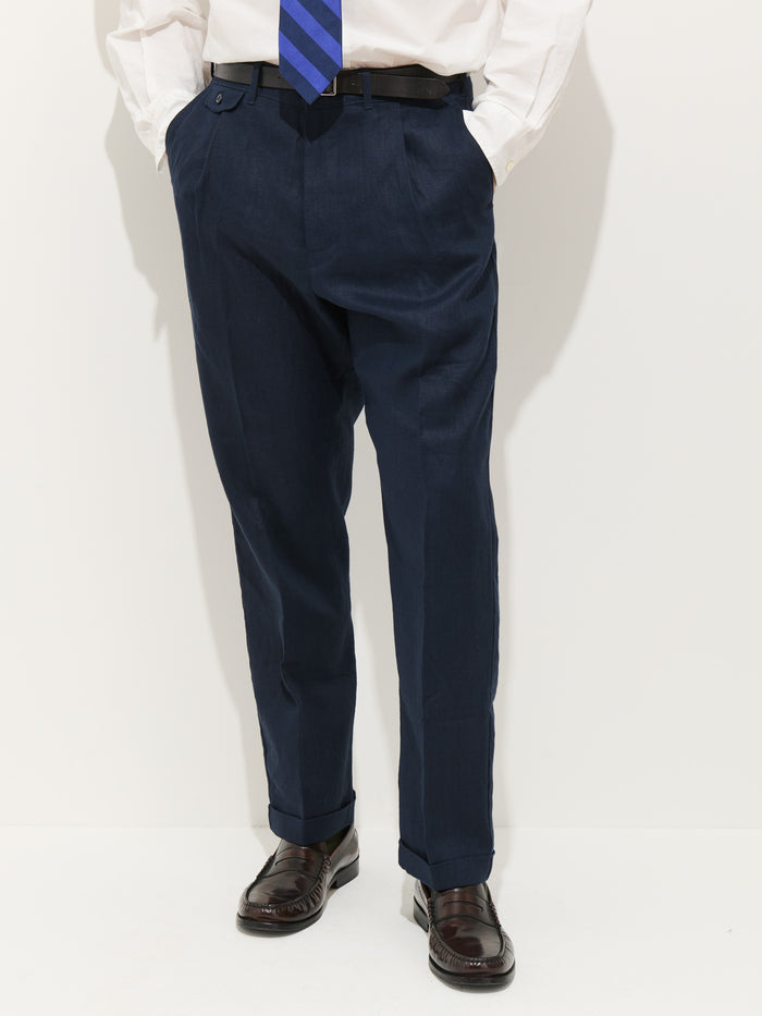 A person wearing the Standard Pleated Pant In Linen (navy), a white dress shirt, black belt, blue striped tie, and dark brown dress shoes stands with hands in pockets against a plain white background.