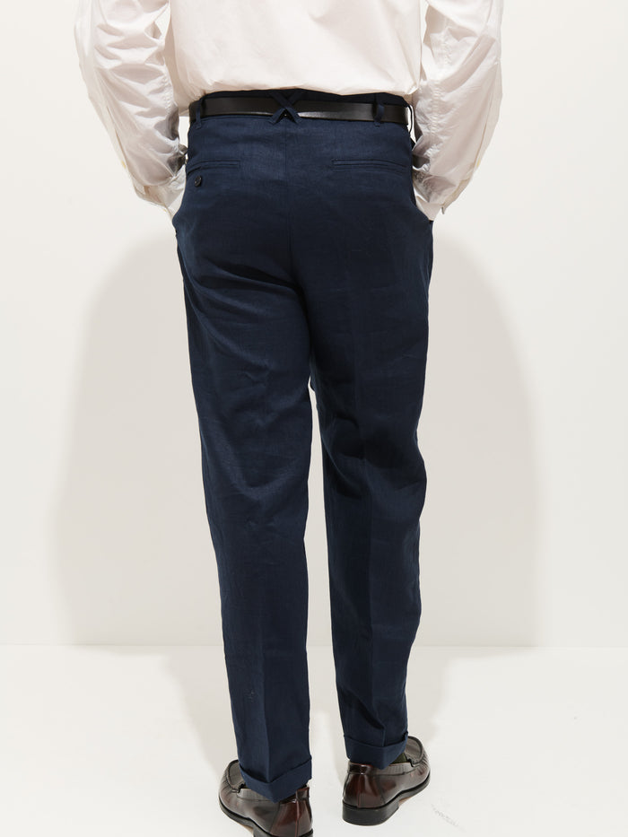 A person stands facing away in a white shirt, dark belt, Standard Pleated Pant In Linen (navy blue), and brown dress shoes against a plain white background.