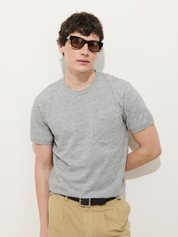 A man wearing dark sunglasses, beige pants, and the Standard Pocket Tee in Slub Cotton in light gray stands against a plain white background with one hand behind his back.