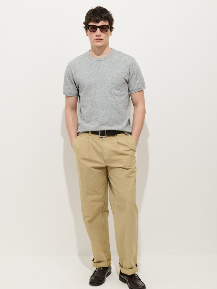 A person stands against a white background wearing sunglasses, the Standard Pocket Tee In Slub Cotton made in LA, beige trousers with a black belt, dark brown shoes, and has hands in pockets.