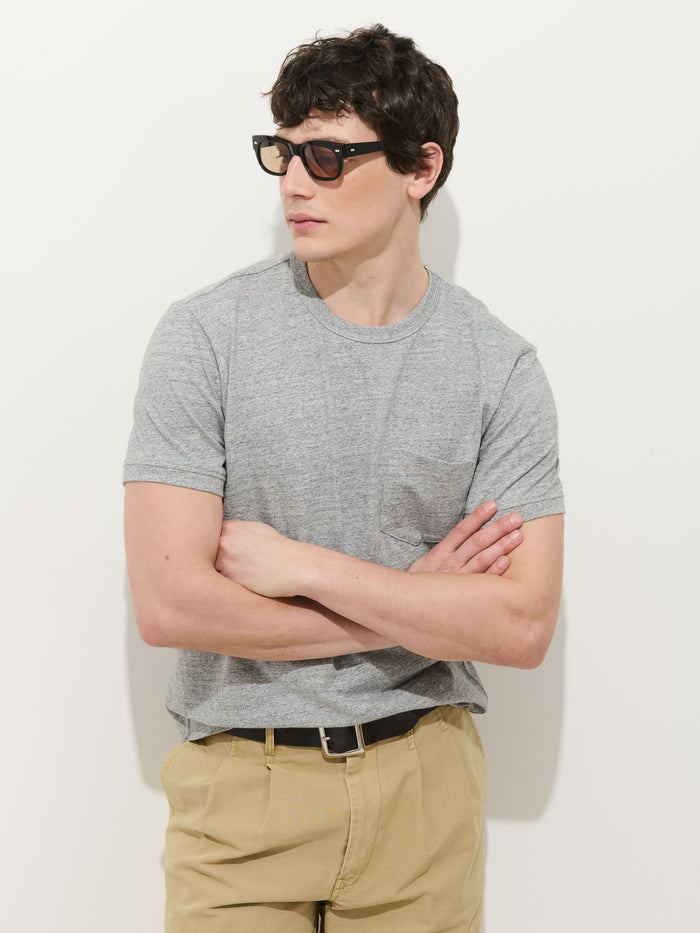 A man stands against a white background with his arms crossed, wearing black sunglasses, beige pants, and the Standard Pocket Tee In Slub Cotton in grey, looking to the side.