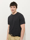 A young man with short, dark hair wears the Standard Slub Pocket Tee in black with beige pants, standing against a plain white background and looking at the camera. Made in LA, this laid-back look features a classic cotton pocket design.