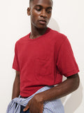 A man wearing the Standard Pocket Tee in Slub Cotton, featuring a chest pocket and a vibrant red hue, looks down and to the side. He has a blue and white striped shirt tied around his waist and stands against a plain white background. Made in LA, this breathable t-shirt combines effortless comfort with laid-back style.