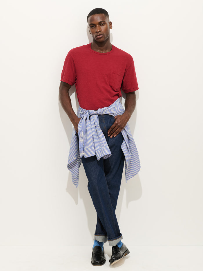 Wearing the Standard Pocket Tee in Slub Cotton in red, paired with dark jeans and black shoes, a person stands against a white wall with a blue-striped shirt tied around their waist, hands in pockets and gazing straight ahead.