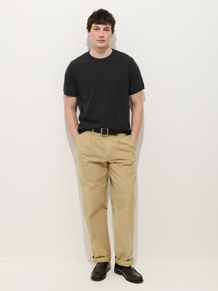 A person stands against a plain white background, wearing the Standard Slub Pocket Tee in black with beige pants, a black belt, and dark brown shoes; hands are in their pockets and they have a neutral expression.