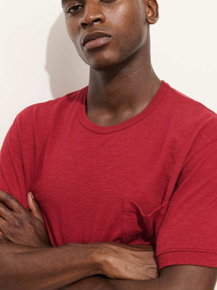 A person stands with arms crossed against a plain white background, wearing the upper half of the Standard Pocket Tee in Slub Cotton—a breathable t-shirt made in LA. Only their upper body and part of their face are visible, highlighting the tee’s signature pocket detail and textured red slub cotton fabric.