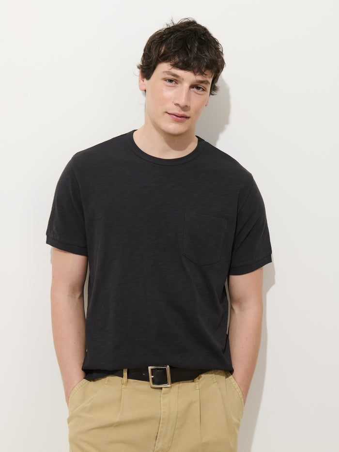 A young man with short dark hair stands against a white background, wearing the Standard Slub Pocket Tee in black, beige pants, and a black belt. His hands are in his pockets as he looks at the camera.