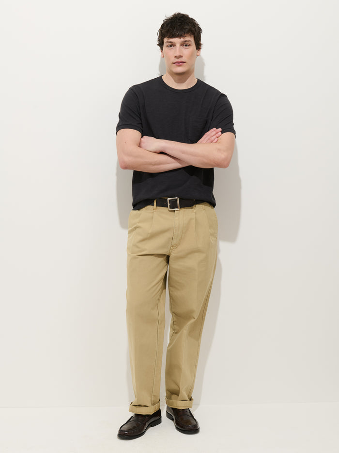 A man with short dark hair and a neutral expression stands against a white background, arms crossed, wearing the Standard Slub Pocket Tee in black, tan khaki pants with a belt, and dark brown shoes.