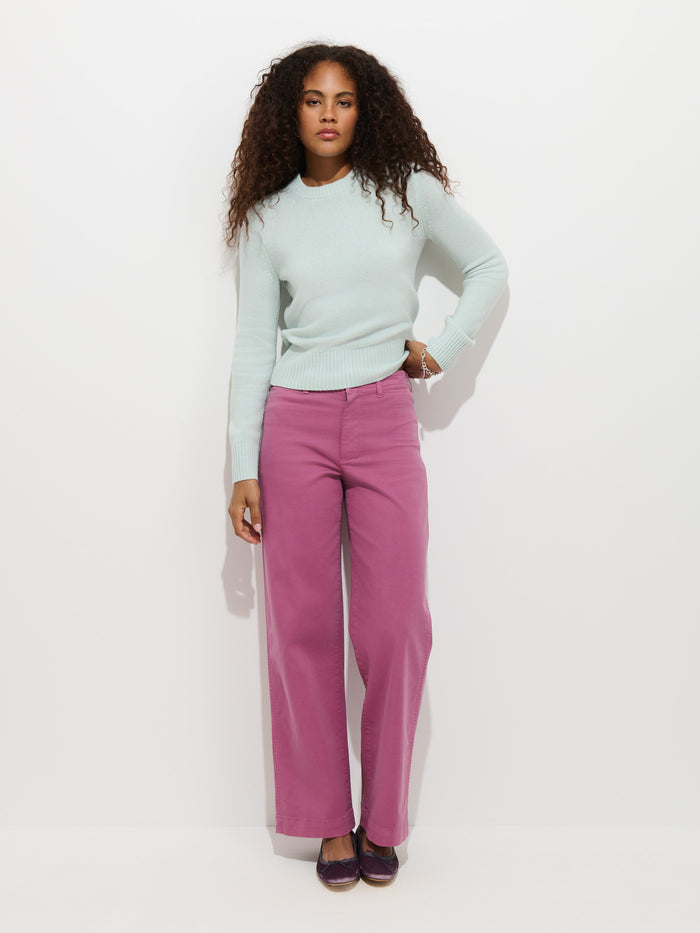 A woman with curly hair stands against a plain white background, wearing a light blue sweater and the Sutton Pant in Vintage Twill—a garment-dyed, high rise, wide-leg chino in pink.