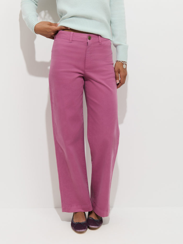 A person stands against a white background, wearing the Sutton Pant in Vintage Twill—a high-rise, wide-leg pink pant garment-dyed for a rich hue—paired with a light blue sweater and purple flats. Only the lower body and one arm are visible.