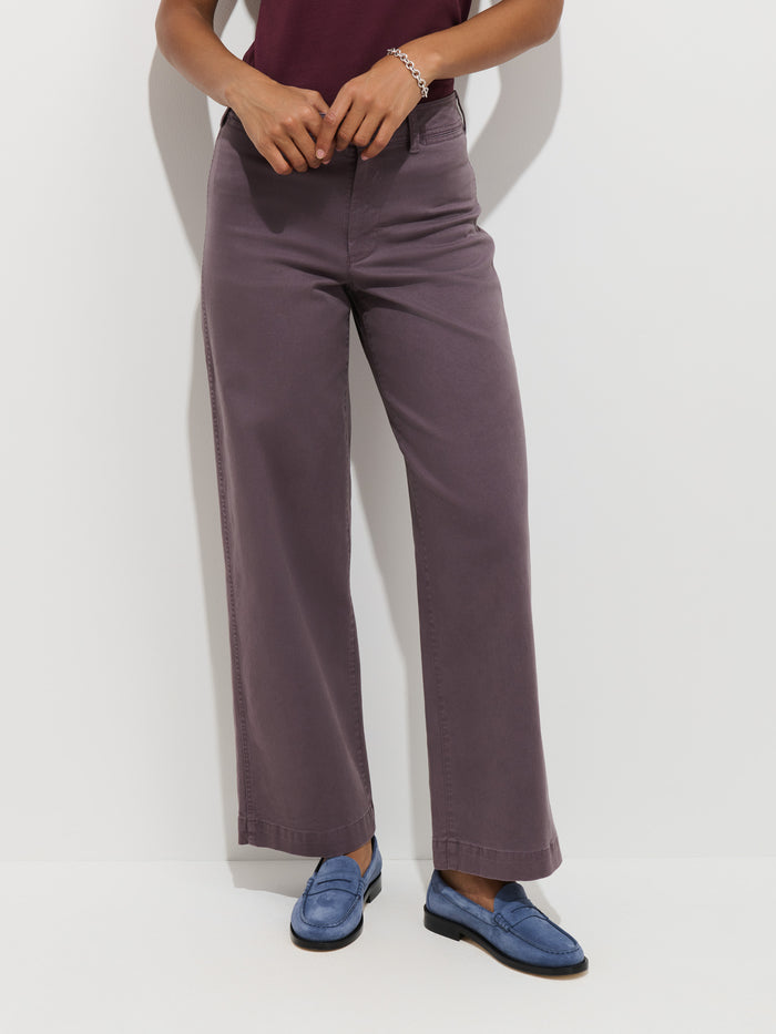 A person wearing the Sutton Pant in Vintage Twill, a wide-leg garment-dyed chino in purple, along with a burgundy top, a bracelet, and light blue loafers stands against a white background.