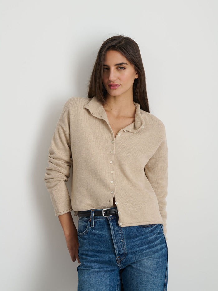 A woman with long brown hair stands against a white wall, smiling slightly with one hand in her pocket. She wears the Taylor Cardigan in Cotton Cashmere over blue jeans and a black belt.