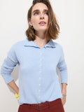 A woman with shoulder-length brown hair wears the Taylor Cardigan in Cotton Cashmere, featuring Troca shell buttons, and rust-colored pants as she stands against a plain white background, looking upward.