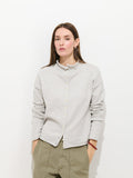 A woman with long brown hair wears the Taylor Cardigan in Cotton Cashmere, styled with olive green pants, standing against a plain white background with her hands in her pockets.