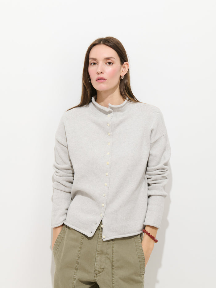 A woman with long brown hair wears the Taylor Cardigan in Cotton Cashmere, styled with olive green pants, standing against a plain white background with her hands in her pockets.