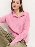 A person with straight brown hair sits against a white wall, calmly looking at the camera while wearing the Taylor Cardigan in Cotton Cashmere, an oversized boxy-fit light pink sweater, paired with olive green cargo pants.