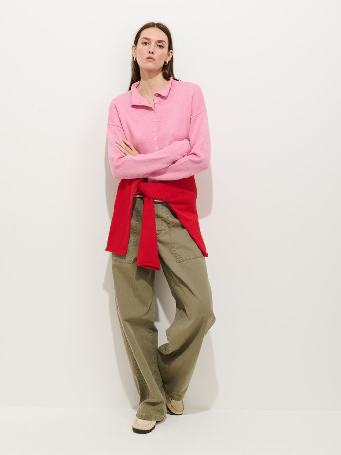 A woman with straight brown hair and gold hoop earrings stands against a white wall, wearing an oversized boxy-fit Taylor Cardigan in Cotton Cashmere (light pink), olive wide-leg pants, beige shoes, and a red sweater tied at her waist.