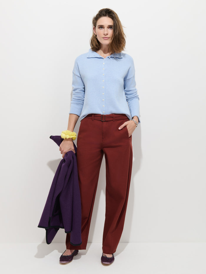A woman stands against a white background, wearing the Taylor Cardigan in Cotton Cashmere in light blue with Troca shell buttons, rust-colored trousers, and purple shoes. She holds a purple coat and has a yellow scrunchie on her wrist.