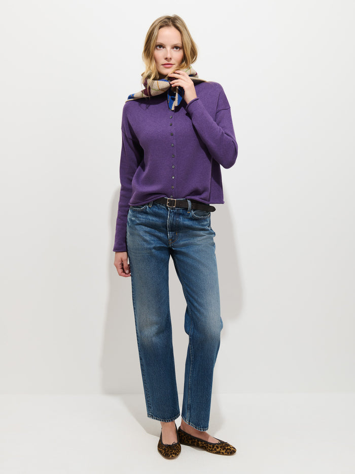A person with blonde hair and a neutral expression stands against a white background wearing the Taylor Cardigan in Cotton Cashmere—an oversized boxy fit purple sweater with Troca shell buttons—paired with blue jeans, leopard print flats, and a colorful scarf around their neck.