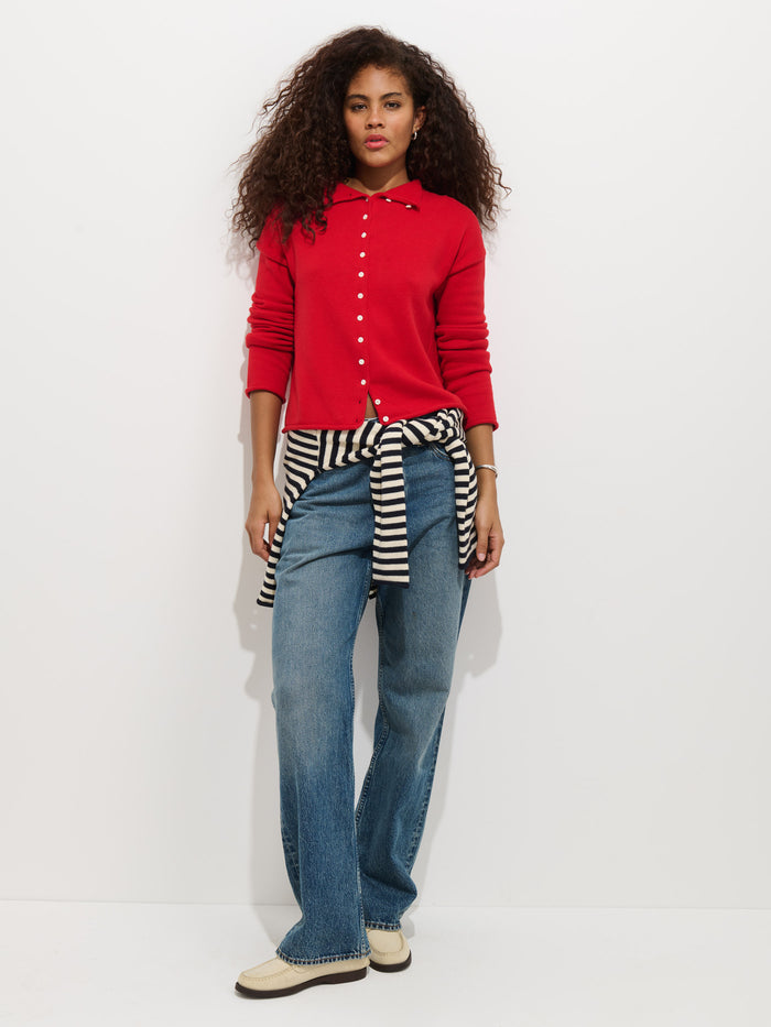 A woman with curly hair wears the Taylor Cardigan In Cotton Cashmere in red, blue wide-leg jeans, and cream loafers. She has a black and white striped sweater tied around her waist and stands against a plain white background.