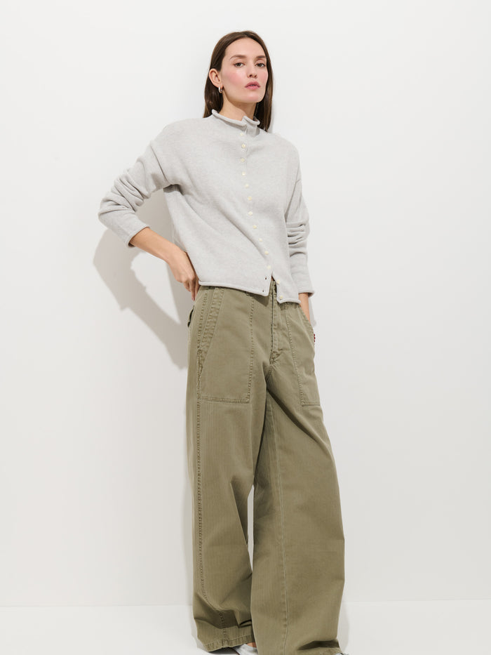 A woman with straight brown hair and a neutral expression stands against a plain white background, wearing the Taylor Cardigan in cotton cashmere and loose-fitting olive green wide-leg pants.