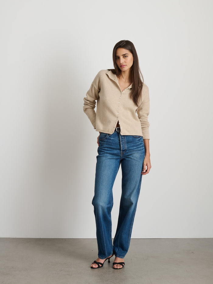 A woman with long brown hair poses confidently against a white wall, wearing the Taylor Cardigan in Cotton Cashmere, paired with blue jeans and black heeled sandals. One hand is in her pocket for a relaxed look.