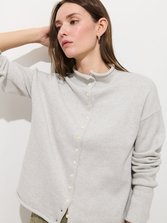 A woman with straight brown hair wears the Taylor Cardigan In Cotton Cashmere, a light gray, boxy oversized sweater with hand-finished construction, paired with light pants. She stands against a white wall, one arm raised and touching her head.