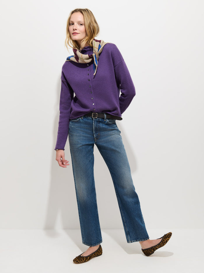 A person in a purple oversized Taylor Cardigan in Cotton Cashmere, blue jeans, and leopard print flats stands against a white background, wearing a patterned scarf around their neck.